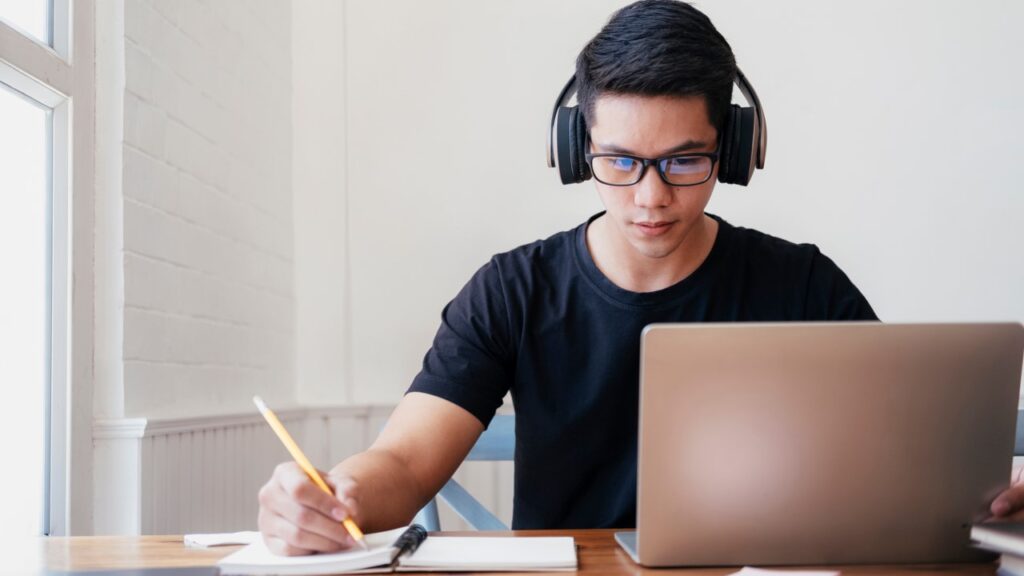 Homem com fone de ouvido, olhando notebook e fazendo anotações em caderno