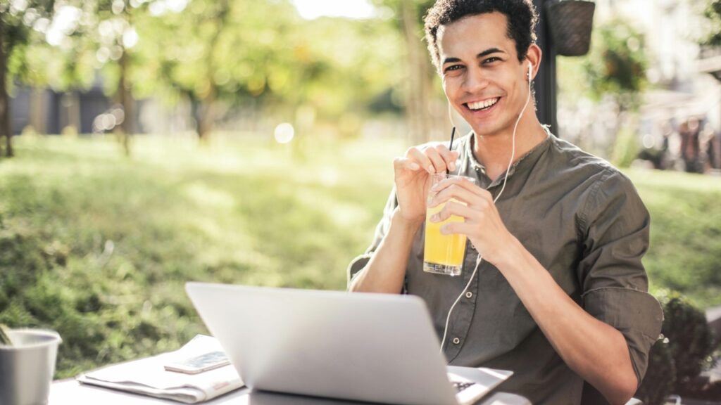 Homem sentado em local aberto com notebook e tomando suco