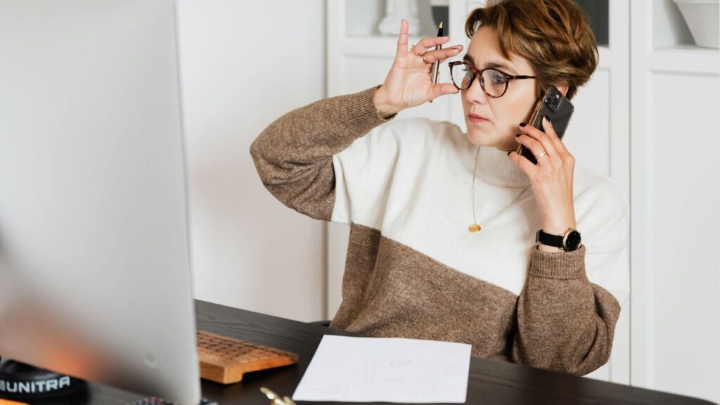 Mulher ajustando óculos enquanto fala ao celular e olha tela de computador